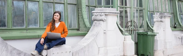 Sonriente joven freelancer en jeans y cárdigan naranja sentado con portátil en la azotea del edificio, pancarta - foto de stock