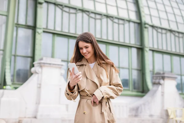 Mujer joven feliz en gabardina mirando el teléfono inteligente cerca del edificio europeo - foto de stock