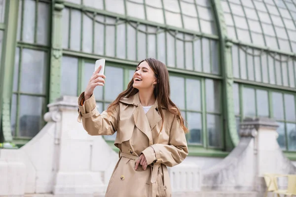 Heureuse jeune femme en trench coat prendre selfie près de bâtiment européen — Stock Photo