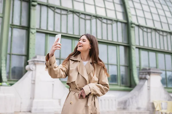 Feliz joven mujer en gabardina tomando selfie cerca de edificio europeo - foto de stock