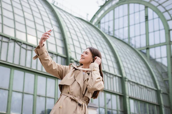 Alegre joven mujer en gabardina tomando selfie cerca de edificio europeo - foto de stock