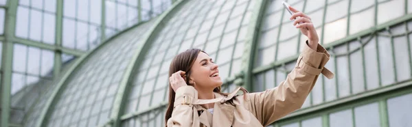 Alegre joven mujer en gabardina tomando selfie cerca de edificio europeo, pancarta - foto de stock