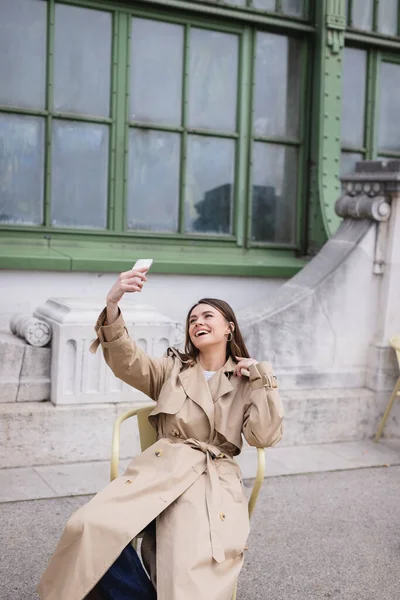 Весела молода жінка в стильному тренч пальто бере селфі біля європейської будівлі — стокове фото