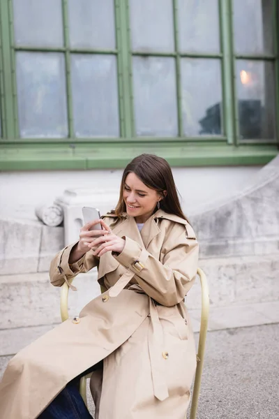 Alegre joven mujer en elegante gabardina usando teléfono inteligente cerca del edificio europeo - foto de stock