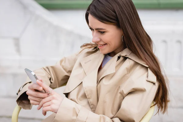 Alegre joven mujer en elegante gabardina usando teléfono inteligente - foto de stock