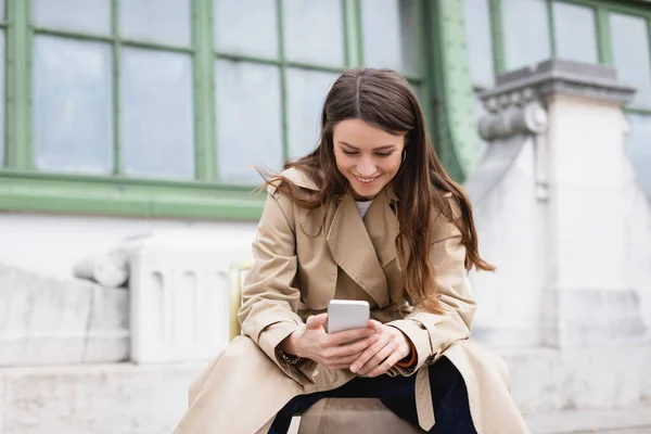 Mujer alegre en gabardina elegante usando el teléfono móvil cerca del edificio europeo - foto de stock