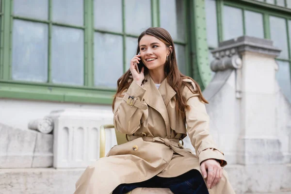 Happy young woman in stylish trench coat having phone call near european building — Stock Photo