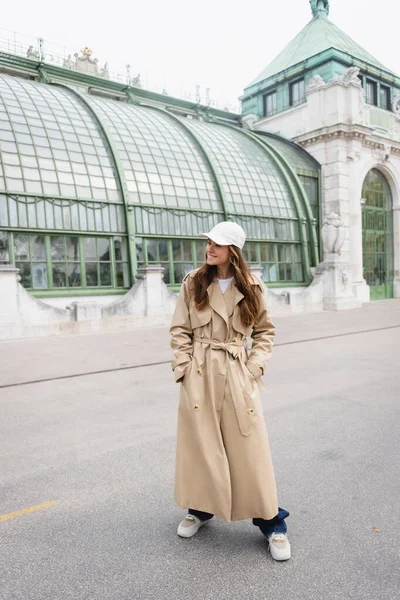 Glückliche junge Frau in Trenchcoat und Baseballkappe, die Hände in den Taschen auf dem Dach eines europäischen Gebäudes stehend — Stockfoto