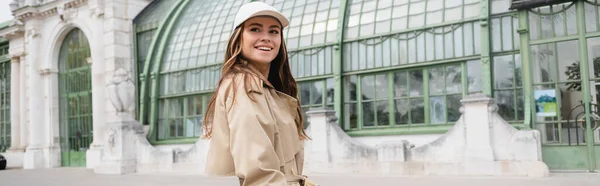 Fröhliche junge Frau in Trenchcoat und Baseballkappe steht auf dem Dach eines europäischen Gebäudes, Banner — Stockfoto