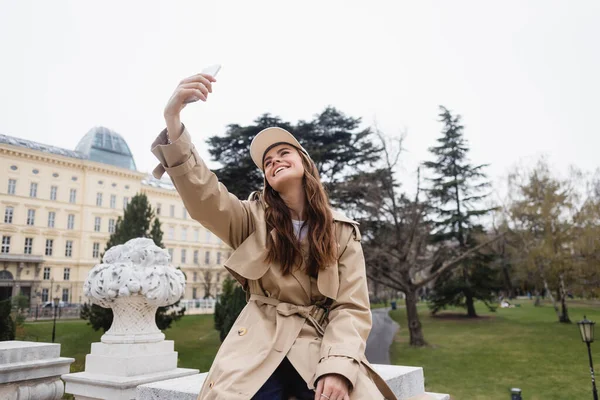 Happy young woman in stylish trench coat and baseball cap taking selfie in european city — Stock Photo