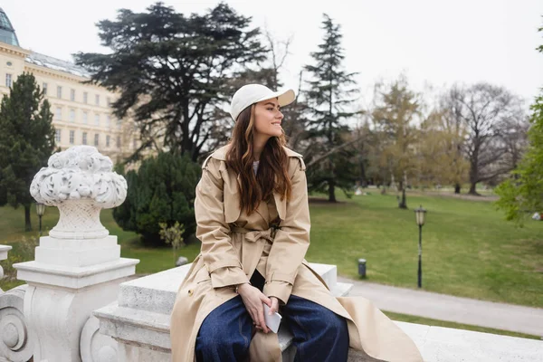 Felice giovane donna in elegante trench e berretto da baseball seduta con smartphone — Foto stock