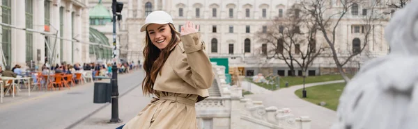 Щаслива молода жінка в стильному траншеї пальто і бейсбольна шапка махає рукою зовні, банер — стокове фото