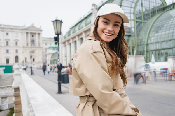 Happy young woman in stylish trench coat and baseball cap — Stock Photo