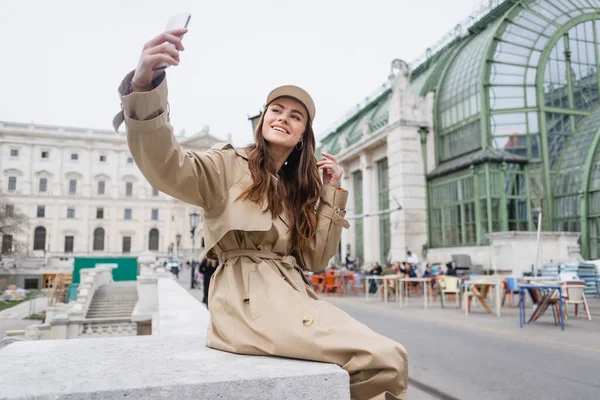 Happy young woman in stylish trench coat and baseball cap taking selfie — Stock Photo