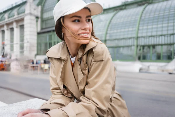 Happy young woman in beige trench coat and baseball cap looking away — Stock Photo
