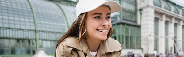 Joyful young woman in beige trench coat and baseball cap looking away, banner — Stock Photo