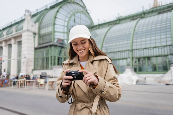 Heureuse jeune femme en trench coat beige et casquette de baseball tenant caméra vintage — Photo de stock