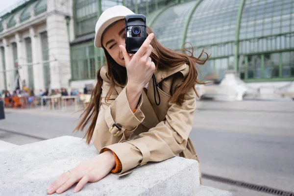 Young woman in trench coat and baseball cap taking photo on vintage camera — Stock Photo
