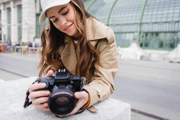 Happy photographer in trench coat and baseball cap looking at digital camera — Stock Photo