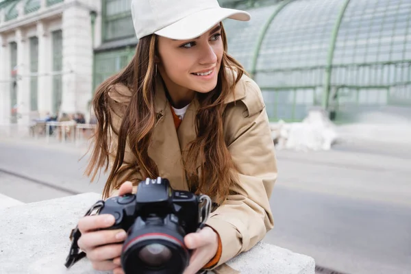 Fotógrafo feliz em casaco de trincheira e boné de beisebol segurando câmera digital — Fotografia de Stock