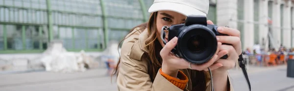 Young photographer in trench coat and baseball cap taking photo on digital camera, banner — Stock Photo
