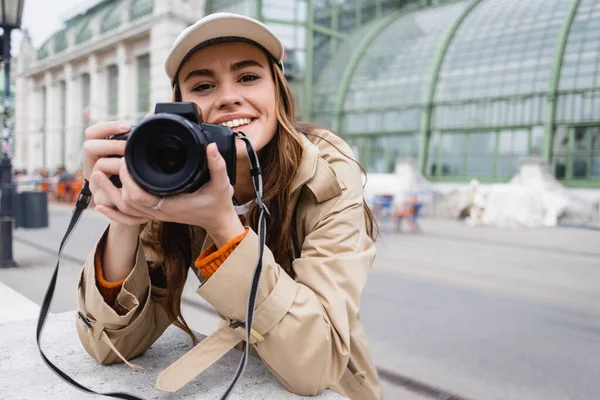Fotógrafo feliz en gabardina y gorra de béisbol tomando fotos en cámara digital - foto de stock