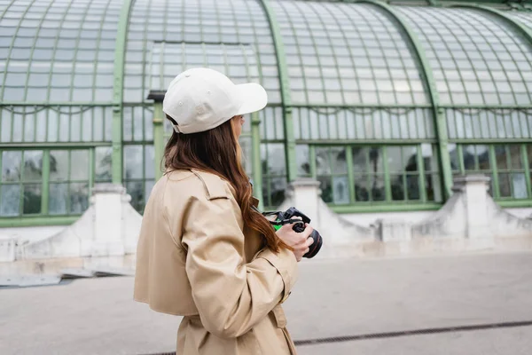 Jeune photographe en trench coat et casquette de baseball tenant un appareil photo numérique dans la ville européenne — Photo de stock
