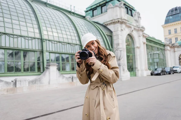 Fotógrafo alegre en gabardina y gorra de béisbol tomando fotos en cámara digital en la ciudad europea - foto de stock