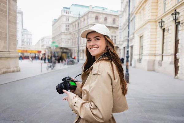 Весела жінка в траншеї пальто і бейсбольна шапка тримає цифрову камеру на вулиці в європейському місті — стокове фото