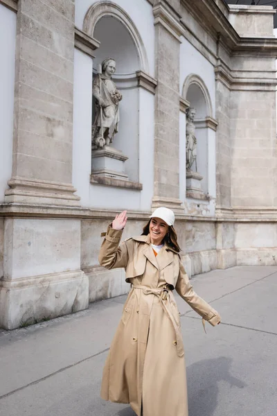 Feliz joven mujer en elegante gabardina y gorra de béisbol caminando y agitando la mano - foto de stock