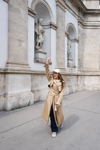 Full length of excited young woman in stylish trench coat and baseball cap walking and waving hand — Stock Photo