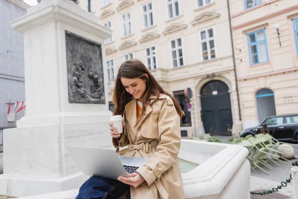 Mujer joven feliz con elegante gabardina mirando a la computadora portátil y sosteniendo la taza de papel - foto de stock