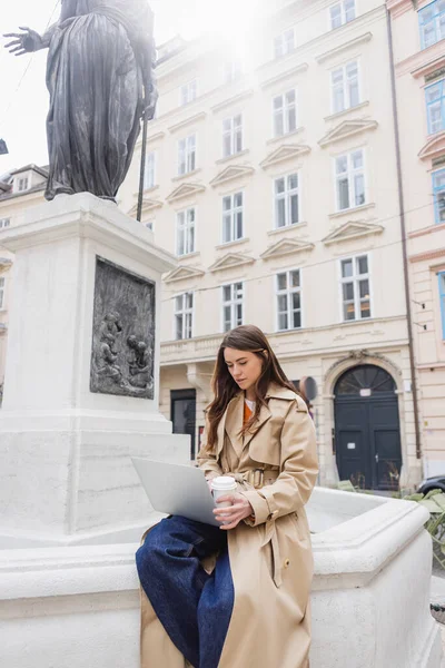 Mujer joven con elegante gabardina mirando a la computadora portátil y sosteniendo la taza de papel en Europa - foto de stock