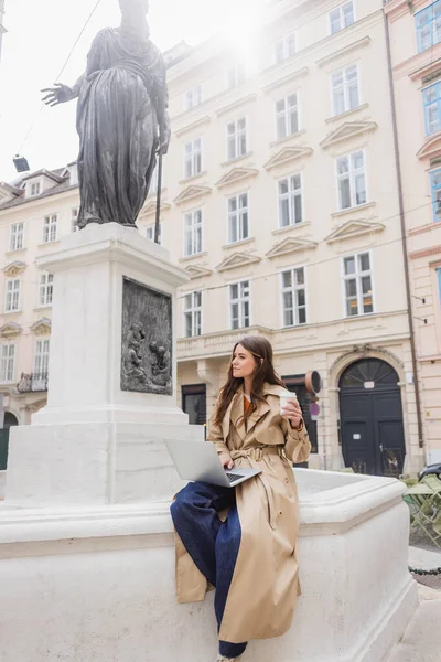 VIENNA, AUSTRIA - MARCH 30, 2022: woman in trench coat sitting with laptop and holding paper cup on european street — Stock Photo