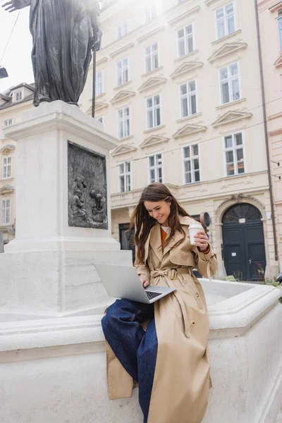 Happy young woman in stylish trench coat looking at laptop and holding paper cup on european street — Stock Photo