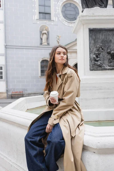 Jeune femme dans élégant trench coat tenant tasse en papier sur la rue européenne — Photo de stock