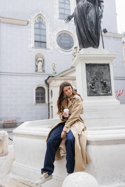 Красива молода жінка в стильному тренч пальто тримає паперову чашку на європейській вулиці — стокове фото