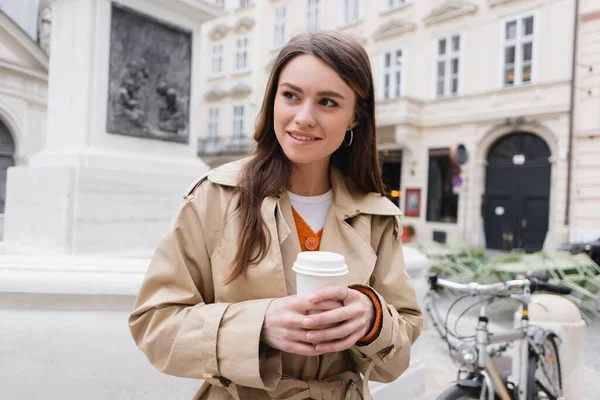 Jeune femme souriante en élégant trench coat tenant tasse en papier sur la rue européenne — Photo de stock