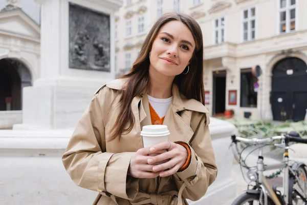 Donna sorridente in elegante trench contenente tazza di carta sulla strada europea — Foto stock