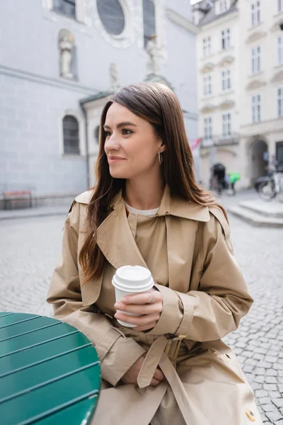 Joven mujer sonriente en elegante gabardina sosteniendo taza de papel y sentado en la terraza de verano en Europa - foto de stock