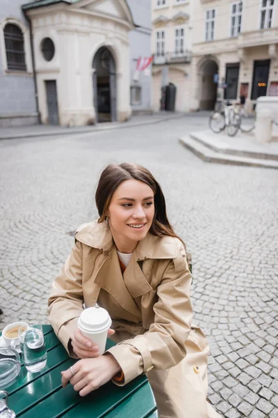 Young pleased woman in trench coat holding paper cup on cafe terrace — Stock Photo