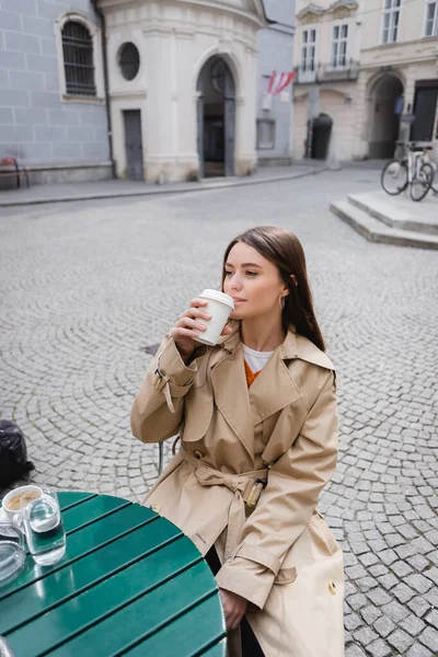 Young woman in trench coat drinking coffee to go on cafe terrace — Stock Photo