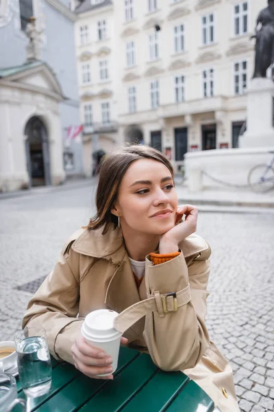 Young woman in trendy trench coat holding coffee to go on cafe terrace — Stock Photo