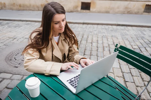Високий кут зору молодої жінки в траншеї пальто, використовуючи ноутбук біля кави, щоб піти — стокове фото