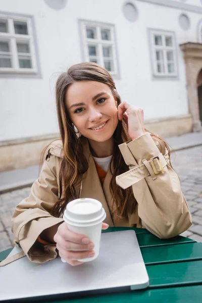 Щаслива молода жінка в модному траншеї пальто тримає каву, щоб піти поблизу ноутбука на літній терасі — стокове фото