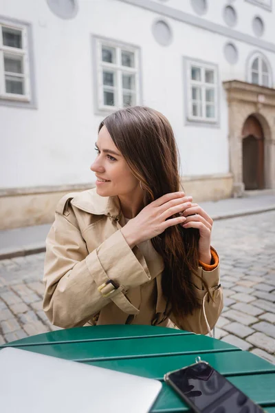 Mujer joven feliz en gabardina de moda ajustar el cabello cerca de la computadora portátil en la terraza de verano - foto de stock