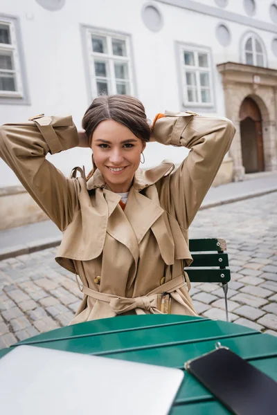 Alegre joven mujer en gabardina de moda ajustar el cabello cerca de la computadora portátil en la terraza de verano - foto de stock