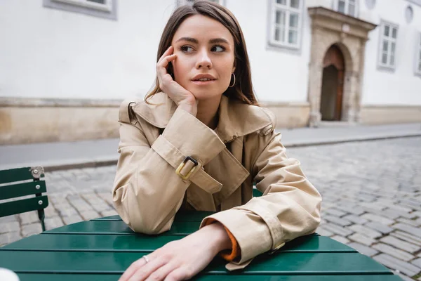 Thoughtful young woman in trendy trench coat sitting in summer terrace — Stock Photo
