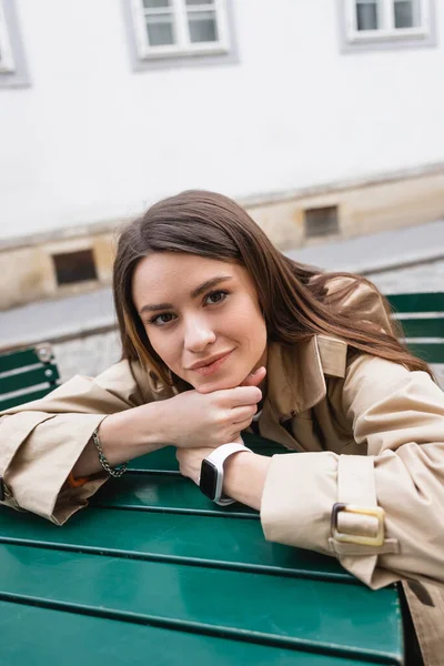 Красива молода жінка в модному траншеї пальто спирається на журнальний столик на літній терасі — стокове фото