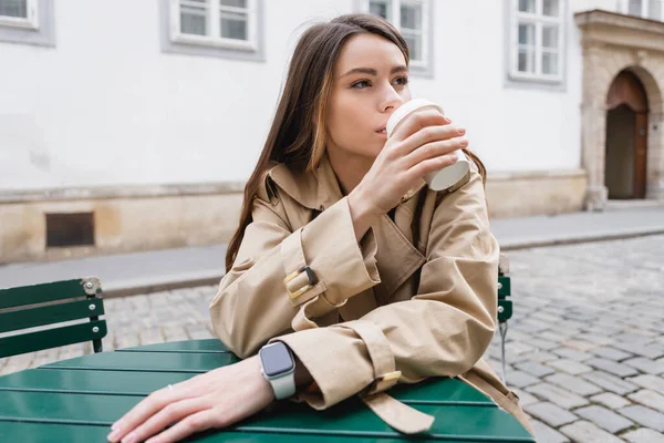 Молода жінка в модному траншеї пальто тримає паперовий стаканчик і п'є каву, щоб піти на літню терасу — стокове фото